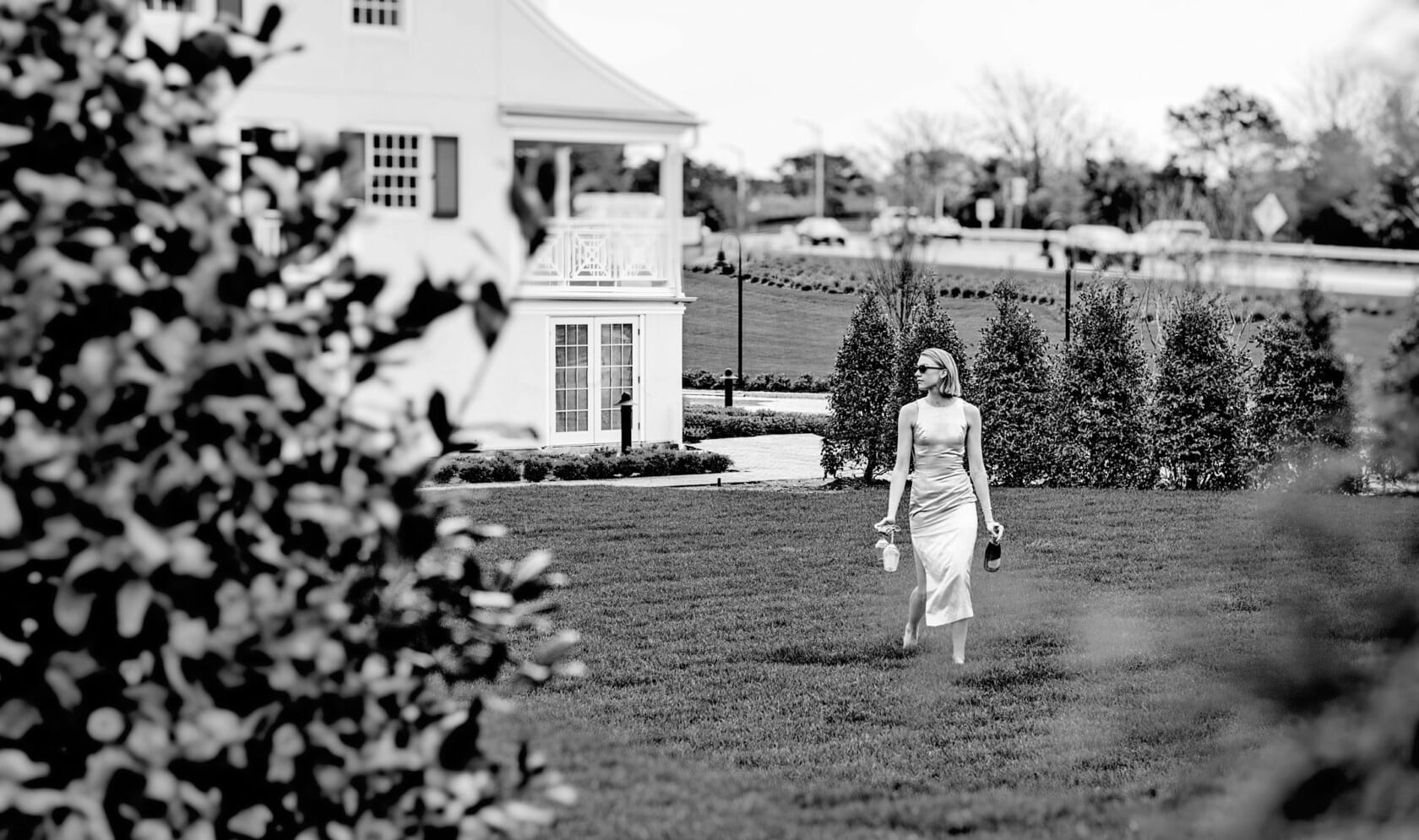 Black and white image of woman crossing garden lawn with historic Canoe Place Inn in the distance.