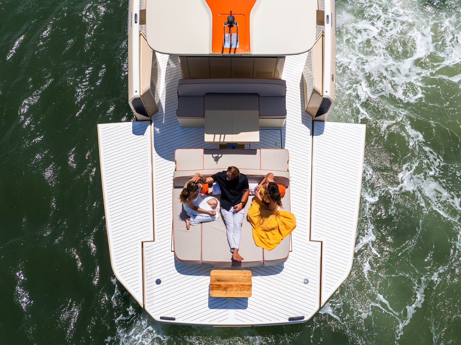 Aerial overhead view of three guests relaxing on the swim platform of a luxury yacht underway on deep green water.